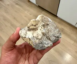 The angular grey-white ambergris piece held in hand indoors, wood floor below