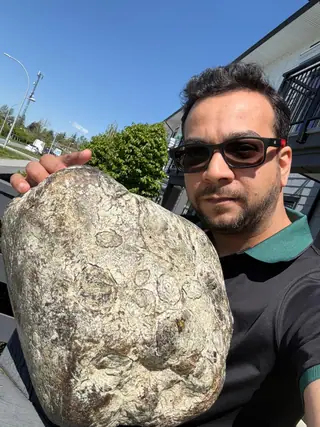 Nabil Valiulla holding the 11.46 kg ringed piece of ambergris up beside his face, blue sky behind